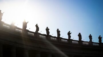 تمثال لأشخاص يقفون على حافة مبنى، silhouetted against السماء الزرقاء. تسطع الشمس من الخلف، مما يخلق تأثير ضوء جميل. يُظهر المشهد العمارة التاريخية والتفاصيل الدقيقة للتماثيل.