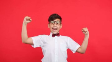 صبي مبتسم يرتدي نظارات وبذلة رسمية مع ربطة عنق، يرفع ذراعيه في وضعية انتصار أمام خلفية حمراء.