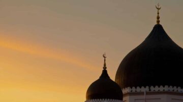 أفق غروب الشمس خلف قباب مسجد مزينة بالهلال، بألوان دافئة تتراوح بين البرتقالي والأصفر، تعكس جمال العمارة الإسلامية في مشهد هادئ.