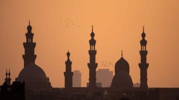 صورة ظلية لمساجد في غروب الشمس، تظهر المآذن والقباب بتفاصيل دقيقة، مع طيور تحلق في السماء. تعكس الألوان الدافئة للغروب جمال العمارة الإسلامية.