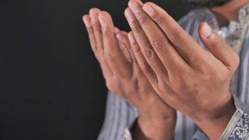 يدان مرفوعتان للصلاة أو الدعاء، تظهران تفاصيل الأصابع والراحة، الخلفية سوداء. تعكس الصورة روحانية العبادة.