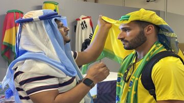 رجل يرتدي غطاء رأس تقليدي يضع قبعة على رأس رجل آخر يرتدي قميص أصفر، في خلفية تحتوي على أعلام متعددة. تعكس الصورة التفاعل الثقافي والاحتفالات.