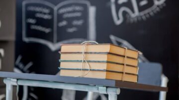 كتب ملونة موضوعة بشكل مرتب على طاولة، مع خلفية تحتوي على سبورة تعكس رسومات تعليمية.