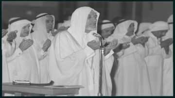 صورة تاريخية لمجموعة من الرجال يرتدون الثوب العربي التقليدي والشماغ، يؤدون الصلاة في مسجد. يظهر أحدهم في المقدمة وهو يدعو، بينما يقف الآخرون خلفه في وضعية الصلاة. الإضاءة خافتة، ما يضفي جواً روحانياً على المشهد.