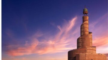 برج مائل بتصميم عصري، يبرز في سماء ملونة بألوان الغروب، مع سحب رقيقة تحيط به، يعكس جمال العمارة الحديثة.