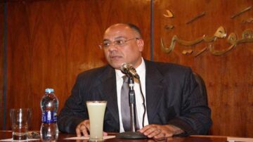 رجل في بدلة رسمية يجلس أمام ميكروفون على طاولة، مع زجاجة ماء وكوب عصير على الطاولة، خلفية خشبية تحمل نصوص باللغة العربية.