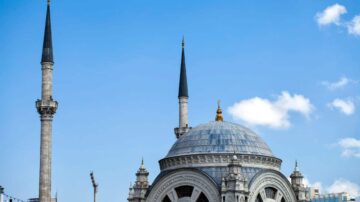 منظر لقمم مسجد تاريخي مع قبتين مميزتين، تتوسطه قبة كبيرة ويحيط به مئذنتان رفيعتان تحت سماء زرقاء صافية مع بعض السحب البيضاء.