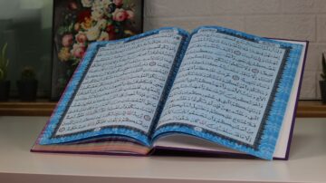 كتاب مفتوح على طاولة، يظهر نصوص باللغة العربية بتصميم ملون. خلفية الصورة تتضمن زهور في إطار مع نباتات صغيرة.