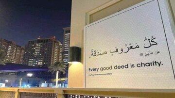 لوحة جدارية في مدينة حديثة تظهر عبارة "كل معروف صدقة" باللغة العربية والإنجليزية، مع خلفية للمباني العالية والإضاءة الليلية. تعكس الرسالة أهمية الأعمال الخيرية في الإسلام.