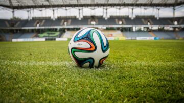 كرة قدم ملونة موضوعة على ملعب أخضر، مع مدرجات فارغة في الخلفية. تصميم الكرة يعكس ألواناً زاهية، مما يبرز أجواء المنافسة الرياضية.