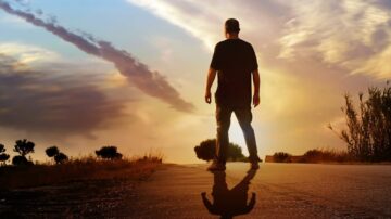 رجل يقف على طريق في وقت الغروب، silhouetted أمام سماء ملونة بسحب جميلة. يشع ضوء الشمس خلفه، مما يخلق ظلالاً طويلة على الأرض.