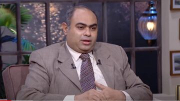 رجل يرتدي بدلة رمادية يجلس في برنامج حواري، يتحدث بجدية مع خلفية تحتوي على نباتات وزخارف داخلية دافئة. الإضاءة خافتة تعكس أجواء مريحة.