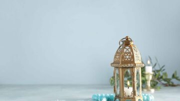 فانوس ذهبي مزخرف مع شمعة مضاءة، موضوع على طاولة مع خرز أزرق وزهور خضراء، خلفية هادئة بلون أزرق فاتح.