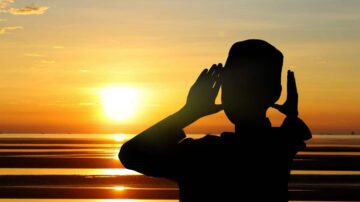 شخص silhouetted أمام غروب الشمس، يضع يديه حول فمه وكأنه ينادي. السماء ملونة بألوان دافئة، والمياه تعكس ضوء الشمس. مشهد يعبر عن التواصل والطبيعة.