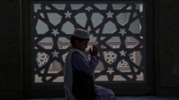 طفل صغير يصلي في المسجد، مع خلفية مزخرفة بنمط هندسي يبرز النجم. الضوء يتسلل من النافذة، مما يخلق جوًا هادئًا وروحيًا.