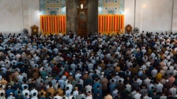 حشود من الرجال يصلون في مسجد كبير، مع وجود أعمدة ملونة في الخلفية. يظهر عدد كبير من المصلين يرتدون ملابس تقليدية، وتفاصيل معمارية مميزة للمسجد.