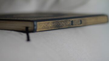 كتاب قديم بجانب حافة فاتحة، يظهر غلافه المزخرف باللونين الذهبي والأزرق، مع حبل أسود مربوط على الجانبين، على سطح أملس.