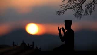 Silhouette of a person praying at sunset with a mosque and tree branch in the background.