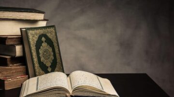 كتاب مفتوح مع نصوص، بجوار مجموعة من الكتب القديمة. يظهر غلاف الكتاب المغلق بلون أخضر مزخرف. الخلفية رمادية، مما يبرز جمال الكتاب.