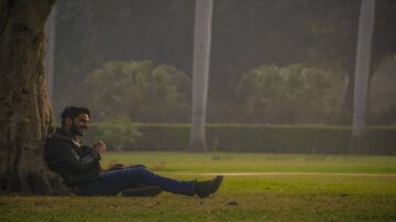 رجل يجلس مستندًا إلى شجرة في حديقة، يبتسم ويحتسي مشروبًا، مع خلفية تحتوي على أشجار ونباتات خضراء في ضوء خافت.