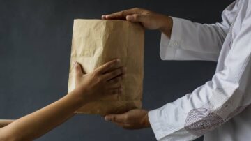 شخص يسلم حقيبة ورقية بنية اللون لشخص آخر، مع التركيز على الأيدي المتداولة للحقيبة. الخلفية داكنة، مما يبرز الفعل البسيط لتبادل الأشياء.