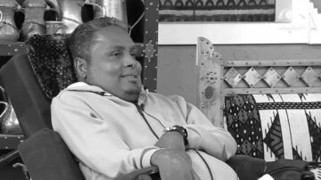 رجل يجلس في كرسي مريح، مبتسم، يرتدي سترة، مع خلفية تحتوي على عناصر تقليدية وزخارف فنية.