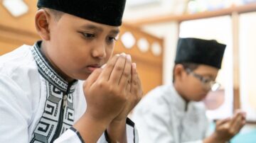 طفلان يرتديان ملابس تقليدية إسلامية، أحدهما يجلس في مقدمة الصورة، معبرًا عن التركيز أثناء الصلاة، بينما الآخر في الخلفية يقوم بالقراءة.