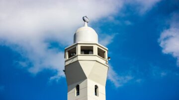 برج مسجد أبيض مع قبة مدورة وقمر في الأعلى، في سماء زرقاء مع بعض الغيوم.