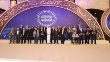 مجموعة من الرجال يقفون على منصة خلال حدث رسمي، يحملون شهادات في أيديهم. خلفهم لوحة مزخرفة تحتوي على نص عربي وزخارف معمارية. الحضور يتضمن شخصًا في wheelchair، مما يعكس تنوع المشاركين.