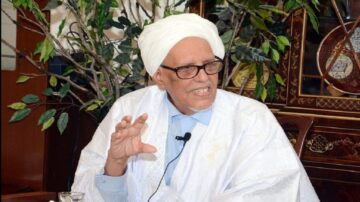 رجل مسن يرتدي عمامة بيضاء وثوب تقليدي، يجلس أمام نباتات خضراء. يعبر عن أفكاره بيديه، ويبدو في محادثة جدية. الخلفية تحتوي على أثاث تقليدي وزخارف.