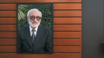 صورة لرجل مسن يرتدي بدلة رسمية مع نظارات وقميص مزخرف، مبتسم أمام خلفية طبيعية وأشجار. الإطار يحيط بالصورة بشكل أنيق على جدار خشبي.