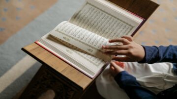 كتاب مفتوح على منضدة مع يد طفل تتصفح الصفحات. النص مكتوب بالخط العربي، ويظهر جزء من الآيات. الخلفية تحتوي على سجادة ملونة.