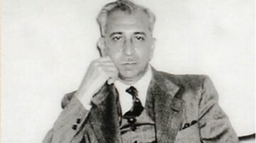 رجل يرتدي بدلة رسمية، يجلس مع ذقنه مستندة إلى يده. تظهر خلفية الصورة باللون الأبيض والأسود، مما يبرز ملامح وجهه وتفاصيل ملابسه.