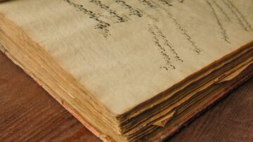 كتاب قديم مع صفحات صفراء ومتآكلة، يظهر نص مكتوب بخط يدوي. الكتاب موضوع على سطح خشبي، يعكس تراث الأدب القديم والكتابة اليدوية.