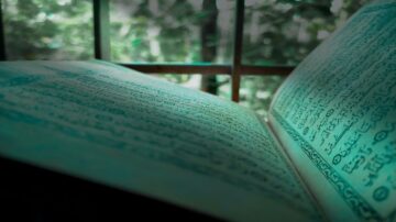 كتاب مفتوح يظهر نصوص دينية بلغة عربية، موضوع بالقرب من نافذة. الضوء الطبيعي يسلط على الصفحات، مع خلفية طبيعية تظهر الأشجار.