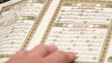 كتاب مقدس مفتوح مع نصوص عربية واضحة، يظهر يد تلمس الصفحات. تتضمن الصفحات آيات مكتوبة بنمط جميل وزخارف ملونة.