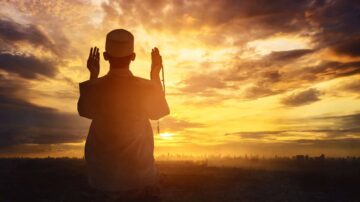 رجل يرتدي عمامة بيضاء يقف في الأفق ويؤدي الصلاة عند الغروب، مع منظر للمدينة في الخلفية، مما يبرز لحظة تأمل روحي.