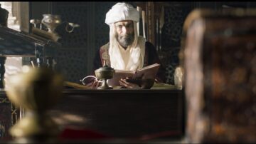 رجل مسن يرتدي ملابس تقليدية عربية يجلس خلف مكتب خشبي، ممسكًا بكتاب مفتوح. يظهر في الخلفية تفاصيل معمارية غنية وأدوات مكتبية قديمة، مما يعكس أجواء تاريخية وثقافية.