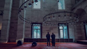 مسجد كبير مع أشخاص يؤدون الصلاة. يظهر في الصورة ثلاثة رجال، اثنان منهم في وضع السجود والآخر واقف، محاطون بأضواء وتفاصيل معمارية تقليدية. الضوء الطبيعي يتسلل من النوافذ، مما يمنح المكان أجواءً هادئة وروحانية.