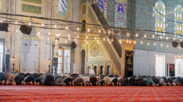 مسجد كبير يضم مجموعة من المصلين يؤدون الصلاة في صفوف متراصة. تظهر الزخارف المعمارية الجميلة والنوافذ الملونة، مع إضاءة خافتة تضفي جواً روحانياً.