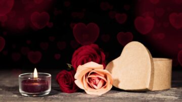 شمعة مضاءة بجانب وردتين، واحدة حمراء والأخرى وردية، مع علبة هدايا على شكل قلب. الخلفية تحتوي على قلوب حمراء، مما يضيف جوًا رومانسيًا ومناسبًا للاحتفالات مثل عيد الحب أو المناسبات الخاصة.
