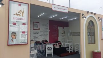 صورة لمعرض يحتوي على جناح للغة العربية. الجناح مزين بشعارات وعناصر بصرية تتعلق بالثقافة العربية، ويظهر فيه مقاعد بيضاء وزخارف فنية. توجد لافتات توضح أنشطة المجتمع المعني باللغة العربية، مع تصميم داخلي ملون.