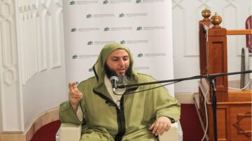 رجل دين يرتدي جلبابًا أخضر ويجلس في مكان مخصص للخطابة، يتحدث من ميكروفون. خلفه جدار مزين بشعارات إسلامية، والبيئة تبدو دينية ومناسبة للوعظ.