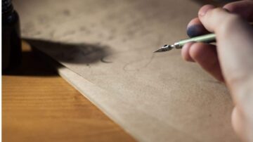 يد تظهر وهي تمسك بقلم حبر على ورقة بنية، مع زجاجة حبر في الخلفية. تصميم فني يبرز الكتابة أو الرسم على الورق.