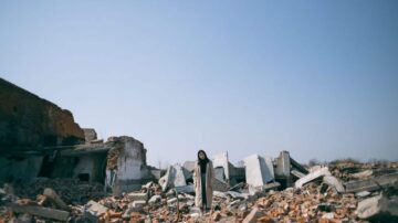 امرأة ترتدي ملابس داكنة واقفة وسط أنقاض مبانٍ مدمرة، مع سماء زرقاء صافية في الخلفية. تظهر الحجارة والخرسانة في حالة فوضى، مما يعكس مشهد دمار.