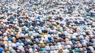 حشود من المصلين يؤدون الصلاة في ساحة واسعة، حيث تظهر رؤوسهم مغطاة بأقمشة ملونة في تنسيق منتظم. تبرز الألوان المتنوعة للمناشف والعمائم، مما يعكس تنوع المشاركين. الأجواء تعكس روحانية الجماعة والعبادة في يوم مقدس.