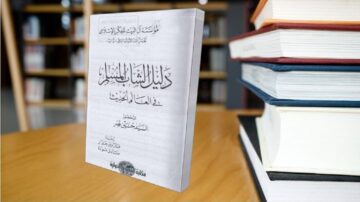 كتاب "دليل المفسر" معروض على طاولة خشبية، يظهر خلفه مجموعة من الكتب الملونة. الكتاب يحمل غلافاً أبيض مع نصوص باللون الأسود، ويبدو أنه يتناول موضوع التفسير.