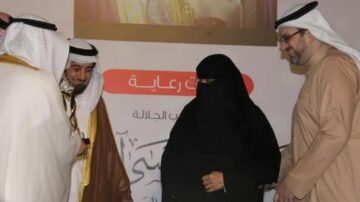 تجمع مجموعة من الرجال والنساء في حدث رسمي، حيث يرتدي الرجال الزي التقليدي العربي، بينما ترتدي المرأة عباءة سوداء تغطي وجهها. يظهر في الخلفية شاشة تعرض معلومات إضافية عن الحدث.