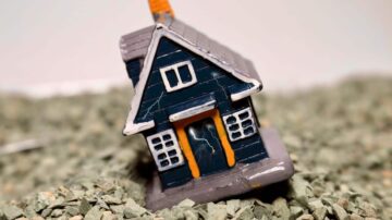 منزل صغير مزخرف باللونين الأزرق والبرتقالي مع تفاصيل واضحة، موضوع على سطح مغطى بالحصى الرمادي. التصميم يظهر cracks في جدران المنزل، مما يضيف لمسة من الواقعية.