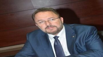 رجل يرتدي بدلة رسمية وزوج من النظارات، يجلس على طاولة، مع خلفية خشبية داكنة. يبدو متفكرًا وجادًا.